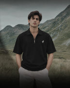 A man wearing a black short-sleeve polo shirt with a small white logo on the chest, standing in a grassy field with mountains in the background.