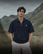 A man wearing a navy blue half-zip polo shirt with a small white embroidered logo on the chest, paired with light beige trousers, standing in a grassy mountain landscape.