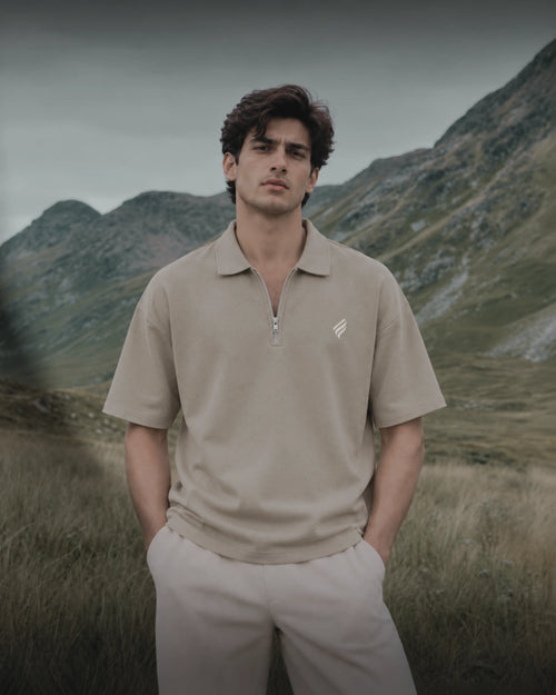 A man wearing a beige short-sleeve polo shirt with a half-zip collar and light beige trousers, standing in a grassy field with mountains in the background.