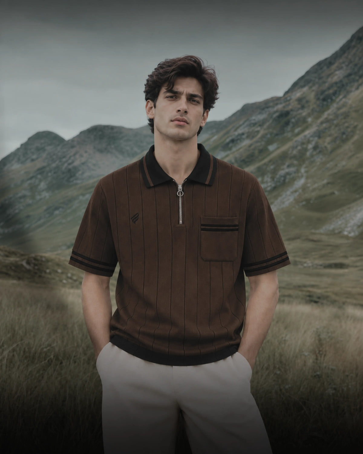 A man wearing a brown short-sleeve polo shirt with a half-zip collar and chest pocket, standing in a grassy mountain landscape.