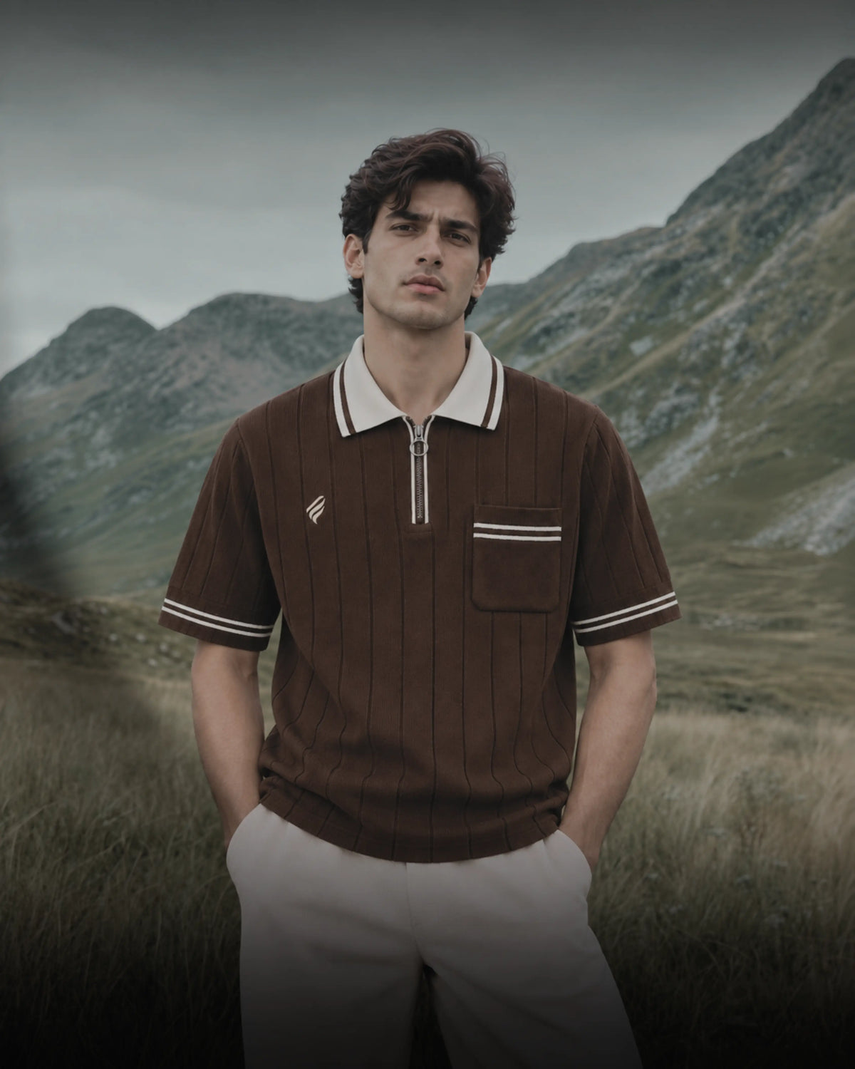 Men’s dark brown ribbed polo shirt with a cream contrast collar, white sleeve stripes, silver quarter zipper, and chest pocket, styled with beige trousers against a mountain landscape.