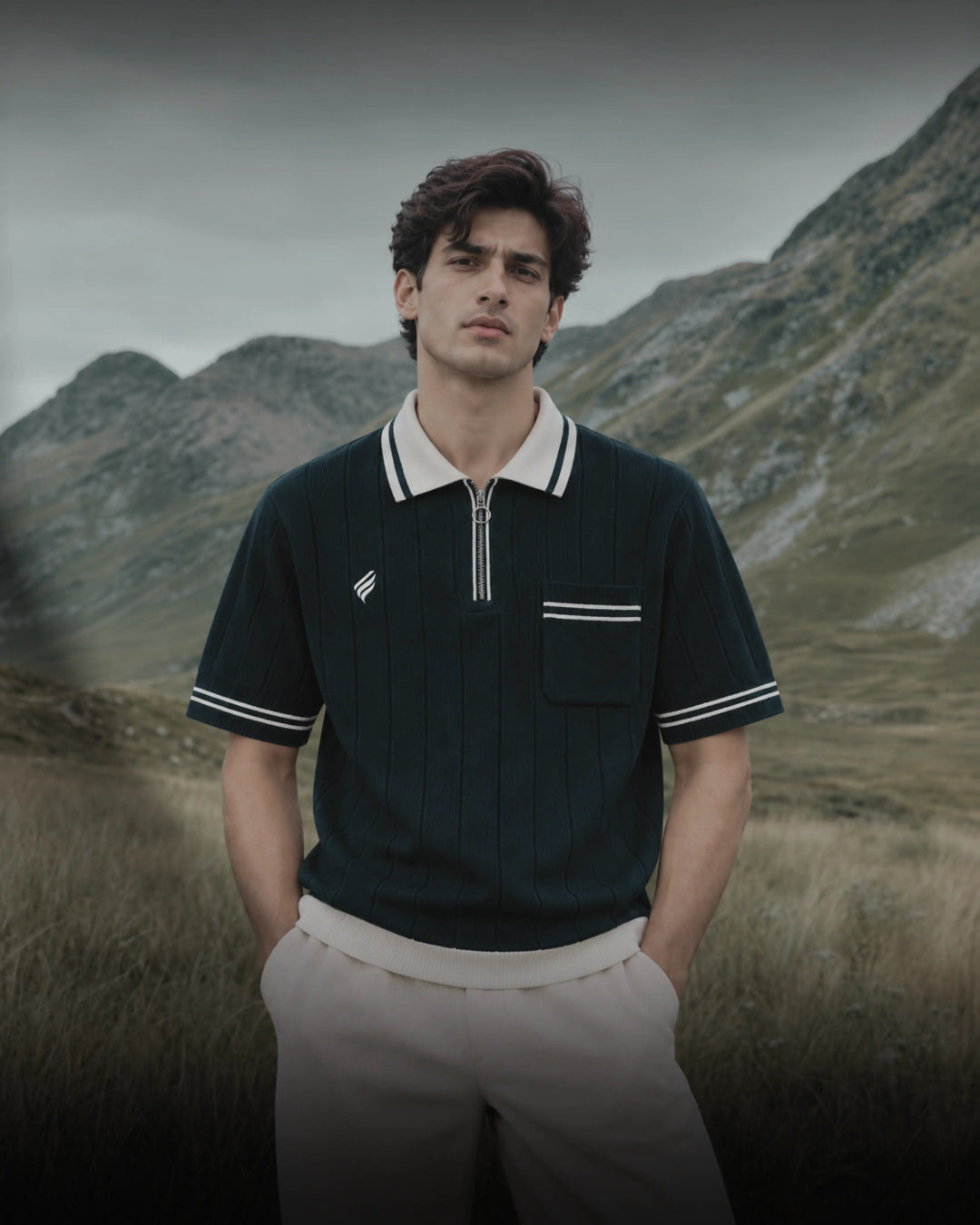 Men’s navy blue ribbed polo shirt with a cream contrast collar, white sleeve stripes, silver quarter zipper, and chest pocket, paired with beige trousers against a scenic mountain backdrop.