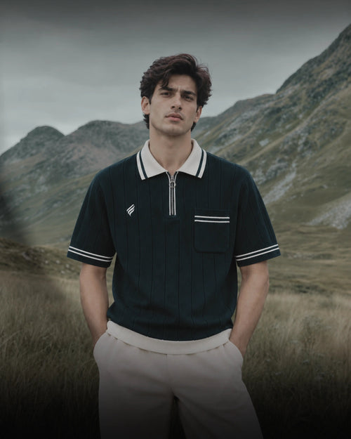 Men’s navy blue ribbed polo shirt with a cream contrast collar, white sleeve stripes, silver quarter zipper, and chest pocket, paired with beige trousers against a scenic mountain backdrop.